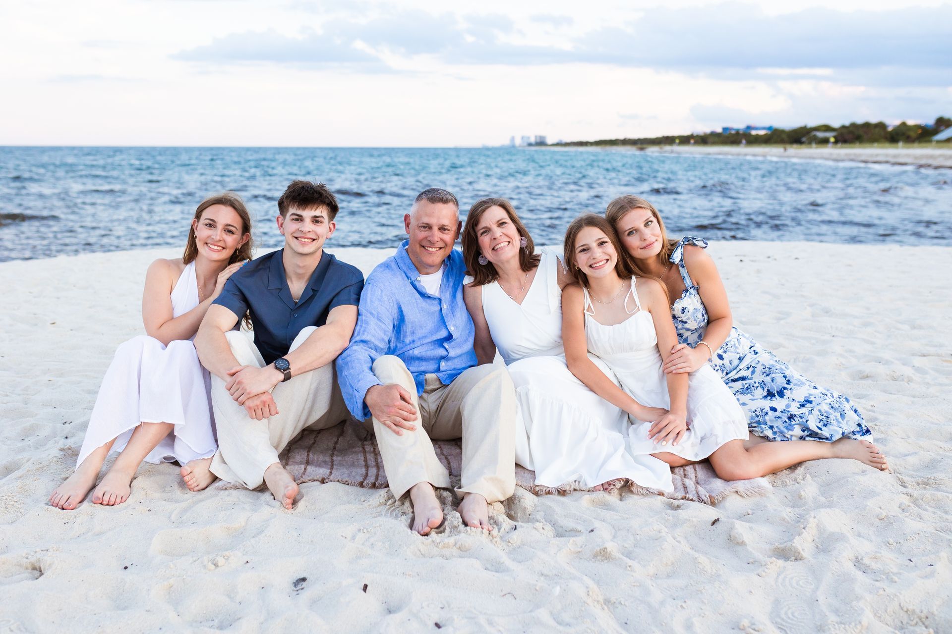 Group of six friends sitting on a white sandy beach by the sea, smiling at the camera.