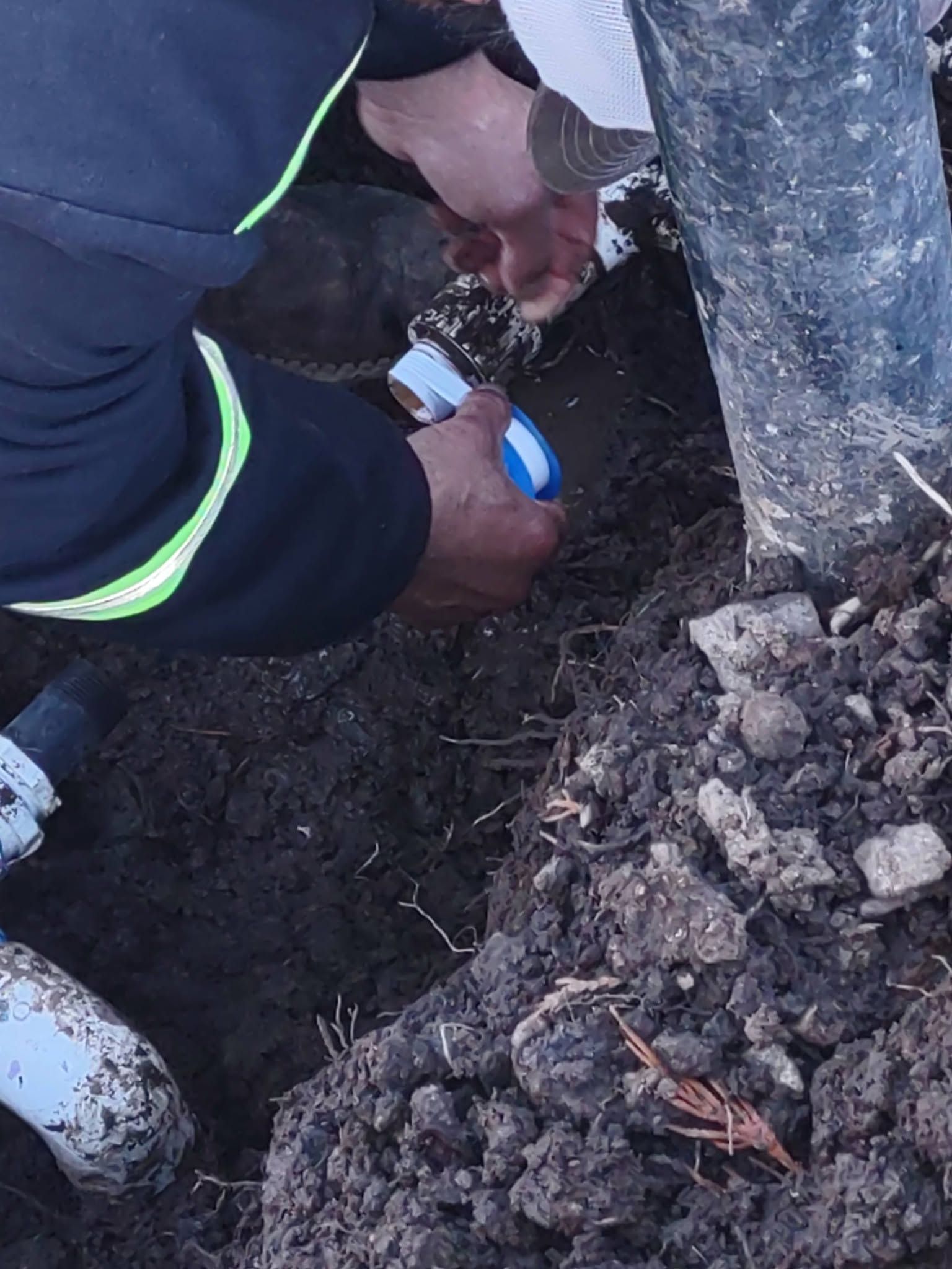 Person in reflective-striped jacket using blue tape to work on a pipe in a dirt trench