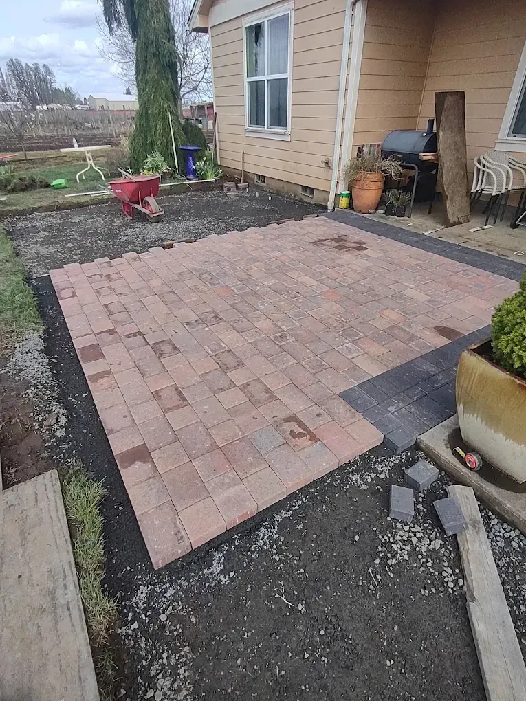 Small brick patio under construction beside a tan house, with gravel and tools around it.
