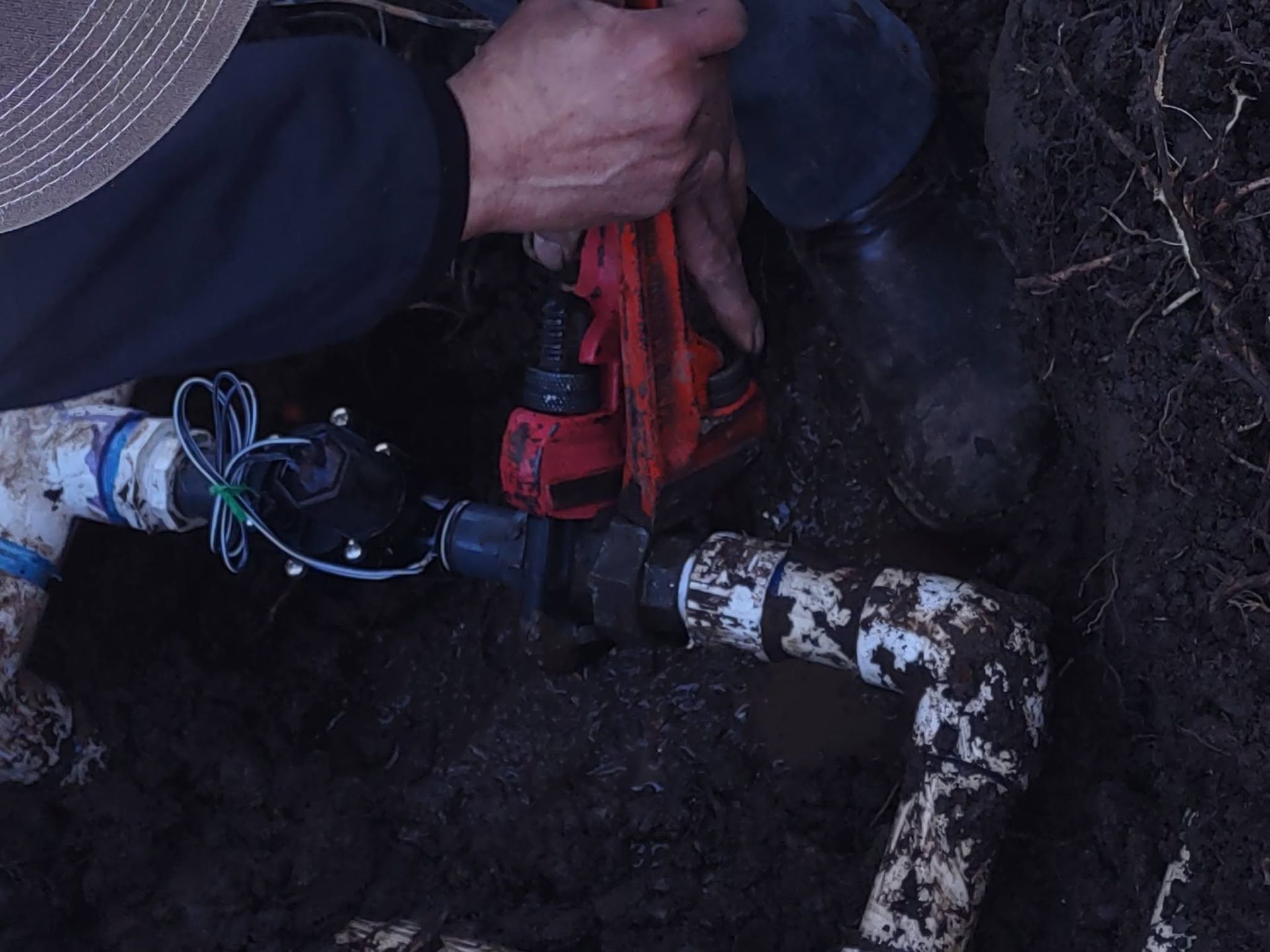 Hands operate a red valve on a muddy pipe in a dark underground utility pit.