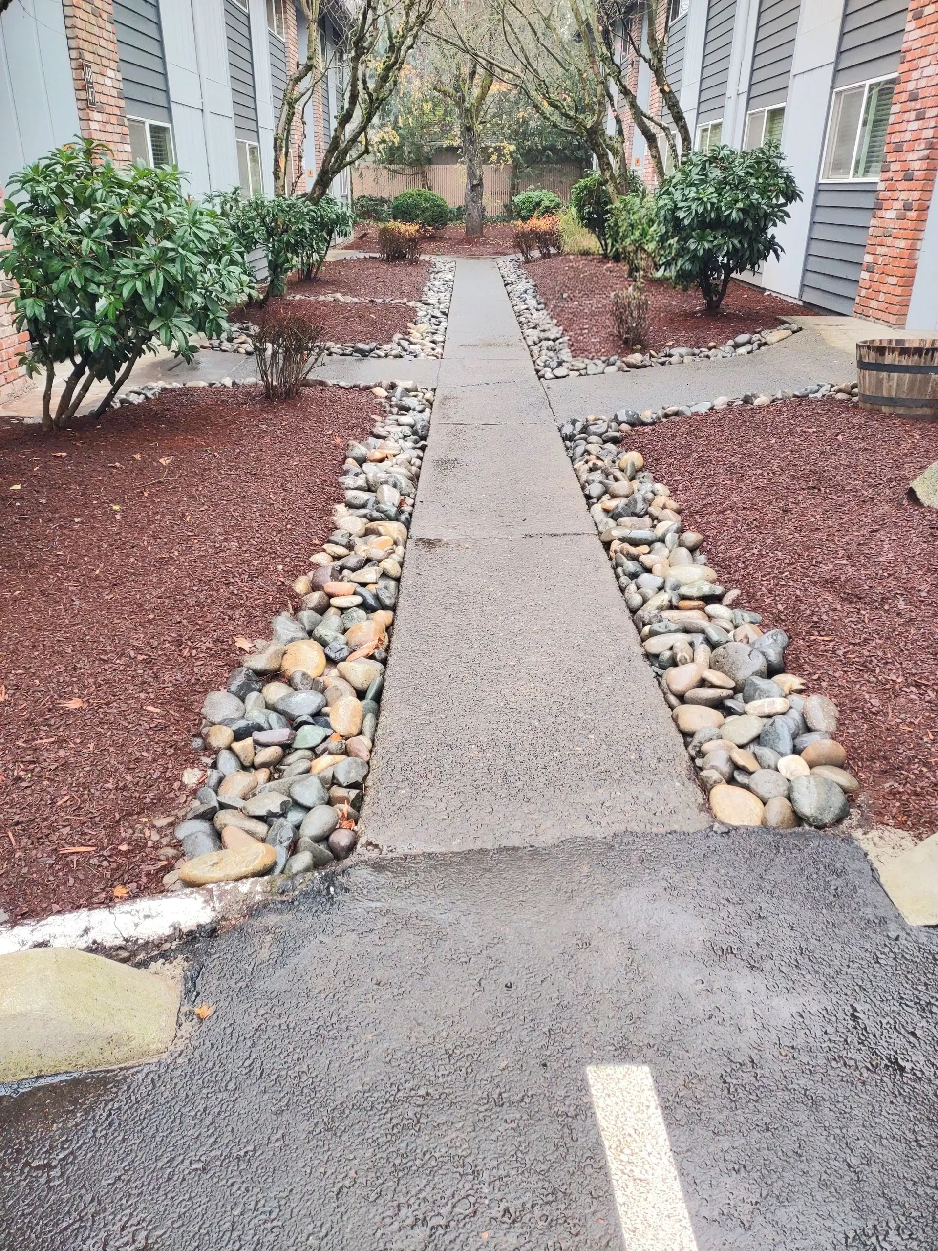 Garden walkway with concrete path, river rocks, and shrubs leading between brick buildings