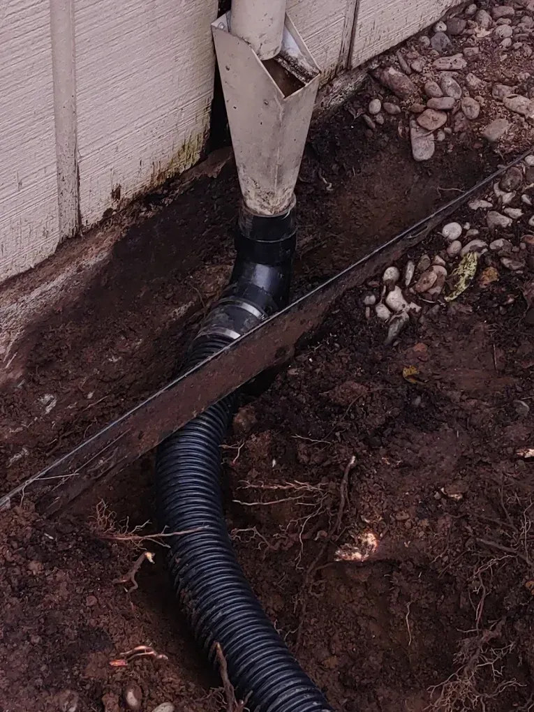 Black drainage pipe running through a dirt trench beside a house foundation, with a broken white downspout above.