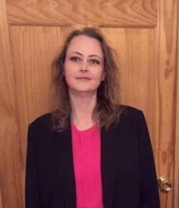 A woman in a pink shirt and black jacket is standing in front of a wooden door.
