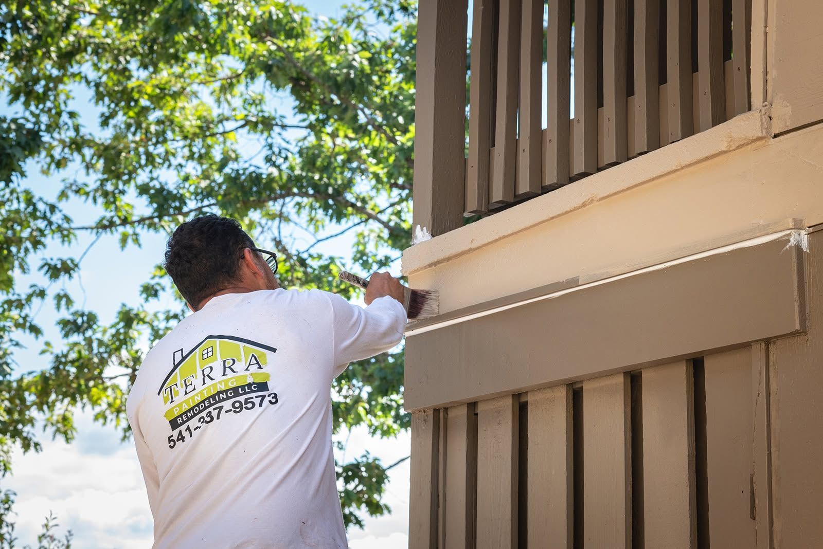 Worker painting the side of a tan house near the roofline under a tree