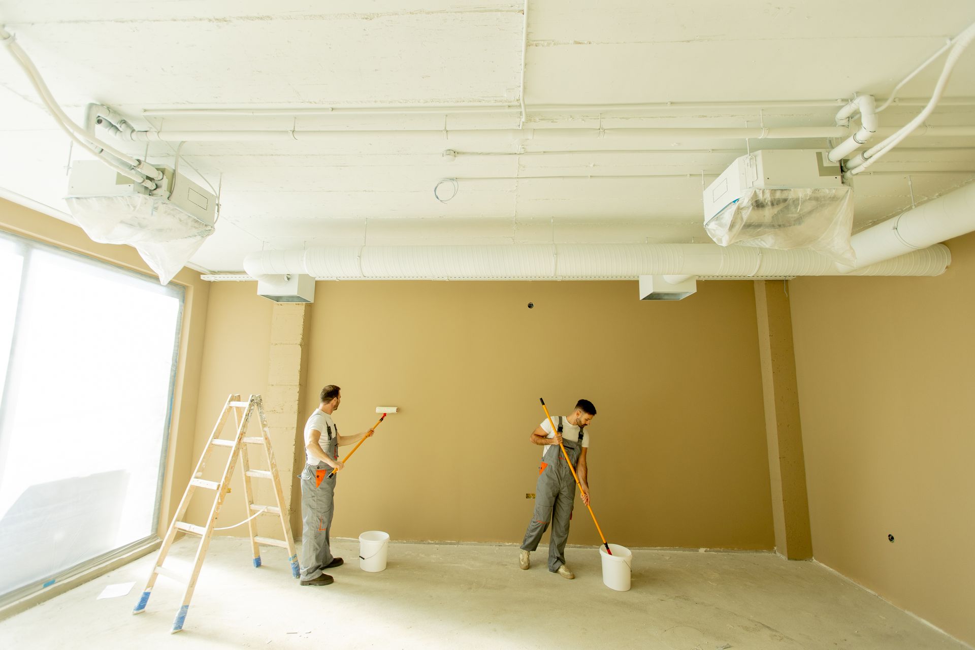 Two painters working on a tan wall inside a bright, unfinished room with ladders and paint buckets.