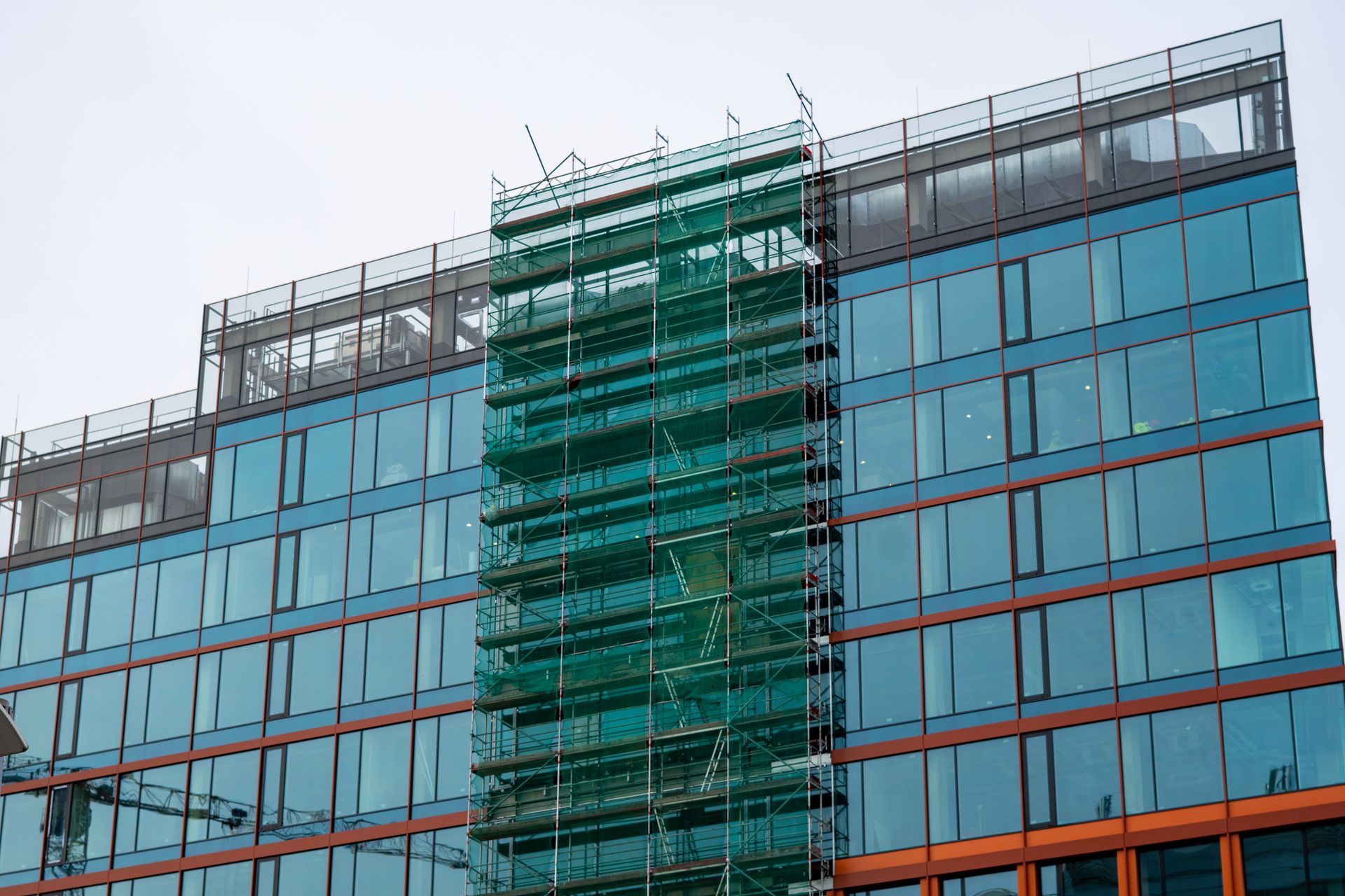 Modern glass office building with green scaffolding and orange trim under an overcast sky