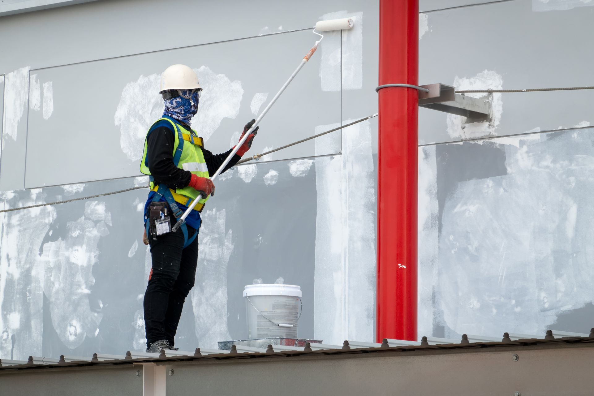 Worker in hard hat painting a building facade from a suspended platform, with a red support beam nearby