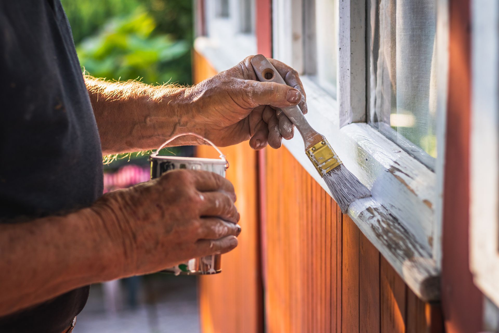 Hands painting a window frame with a brush, holding a paint can beside an orange exterior wall.