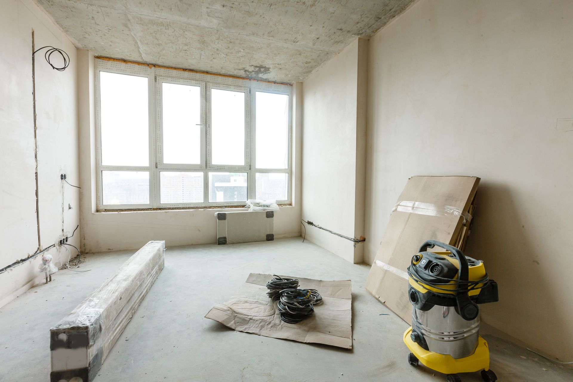 Renovation room with bare walls, large window, paint supplies, and a vacuum cleaner on the floor