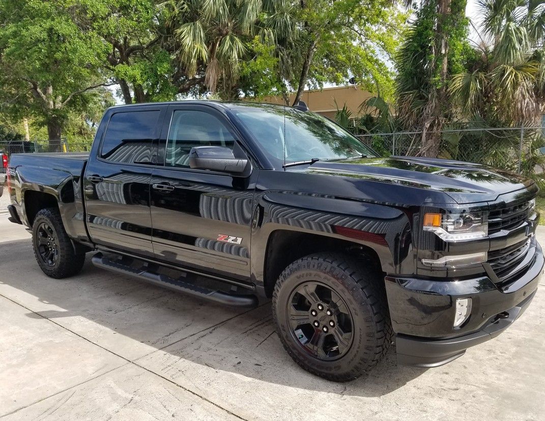 Black pickup truck parked in a driveway beside trees and a chain-link fence.