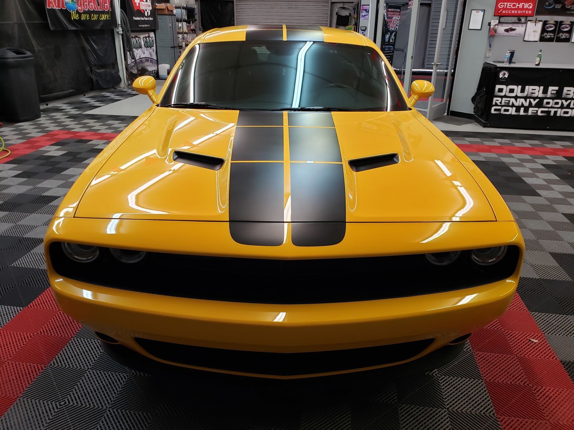A yellow dodge challenger with black stripes on the hood is parked in a garage.