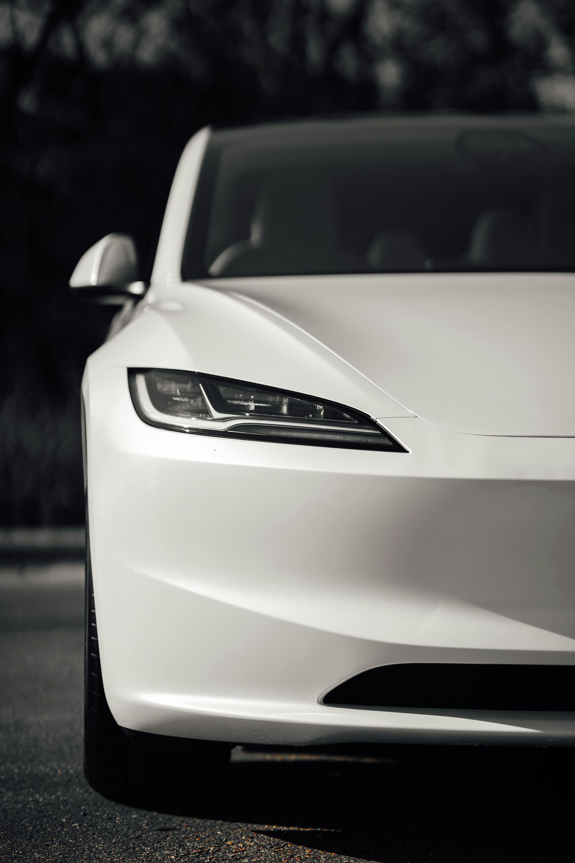 A sleek white Tesla Model 3 sedan in a low-angle, close-up shot focusing on the headlight and hood profile.