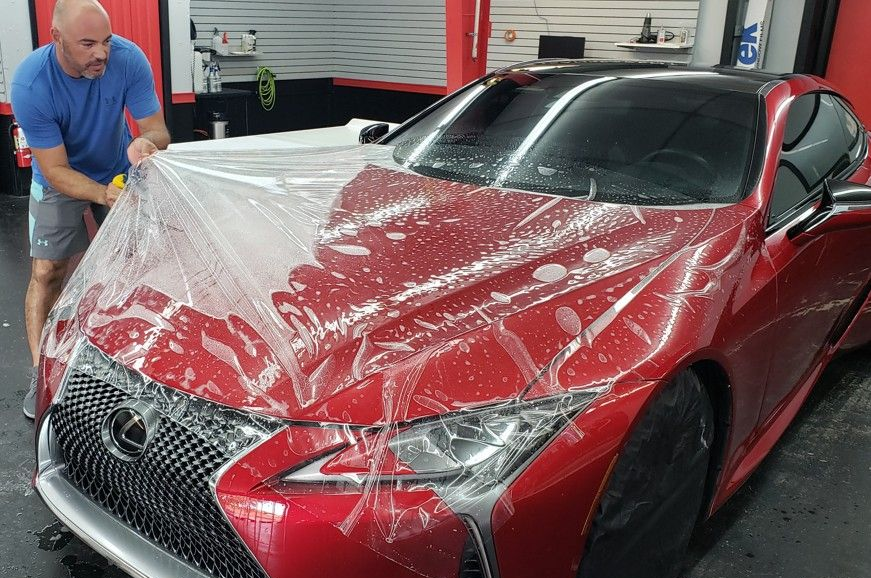Red sports car being wrapped in clear protective film inside a garage by a worker