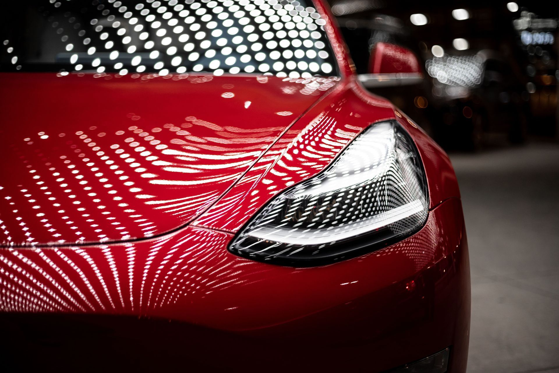 Close-up of a glossy red car hood and headlight under bright, patterned studio lights.