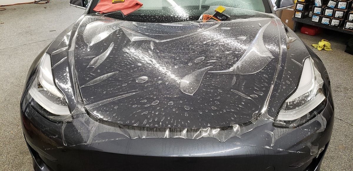A dark grey Tesla with a protective clear film partially applied to its hood inside a garage.