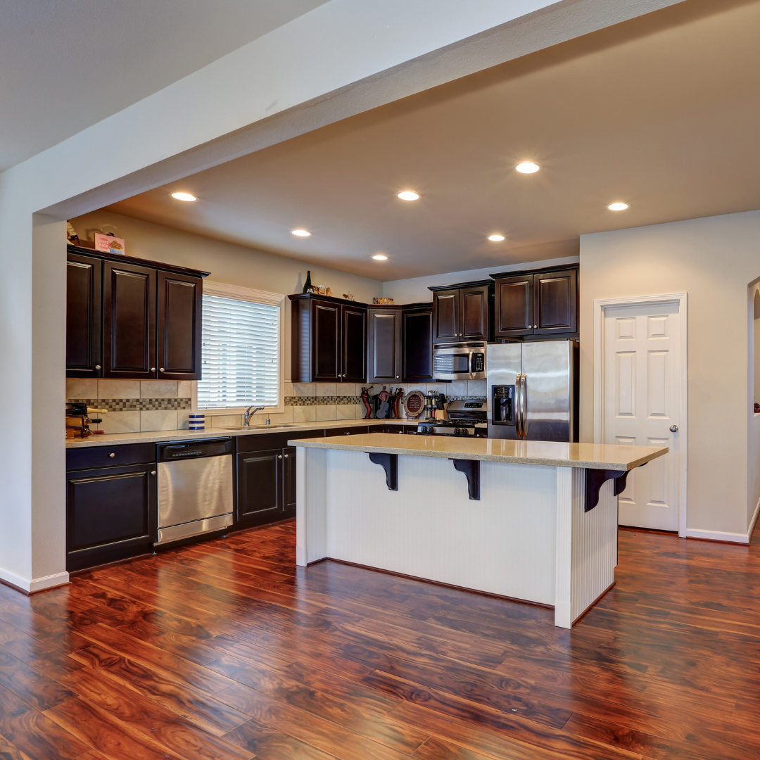 A kitchen with hardwood floors , black cabinets , stainless steel appliances and a large island.