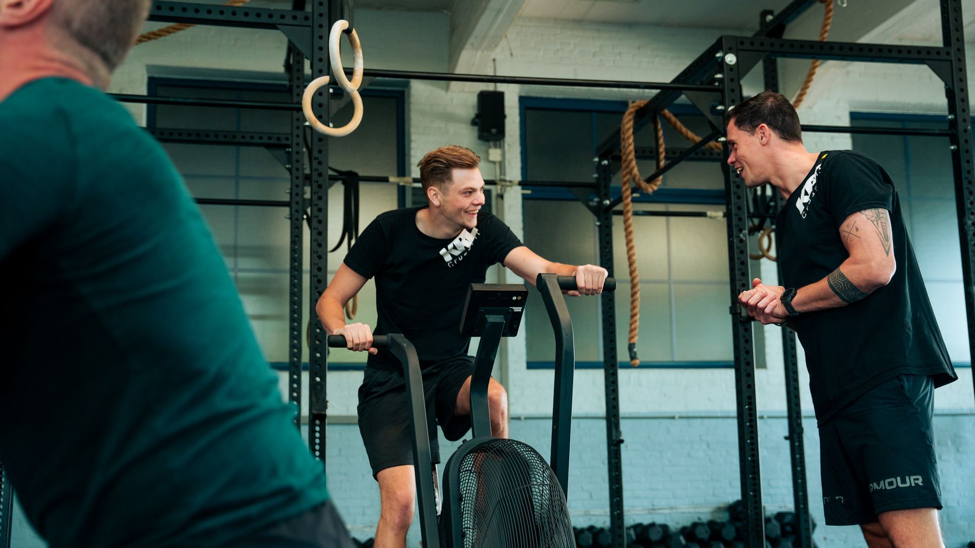 Twee mannen praten met elkaar in een sportschool terwijl een van hen op een hometrainer zit.