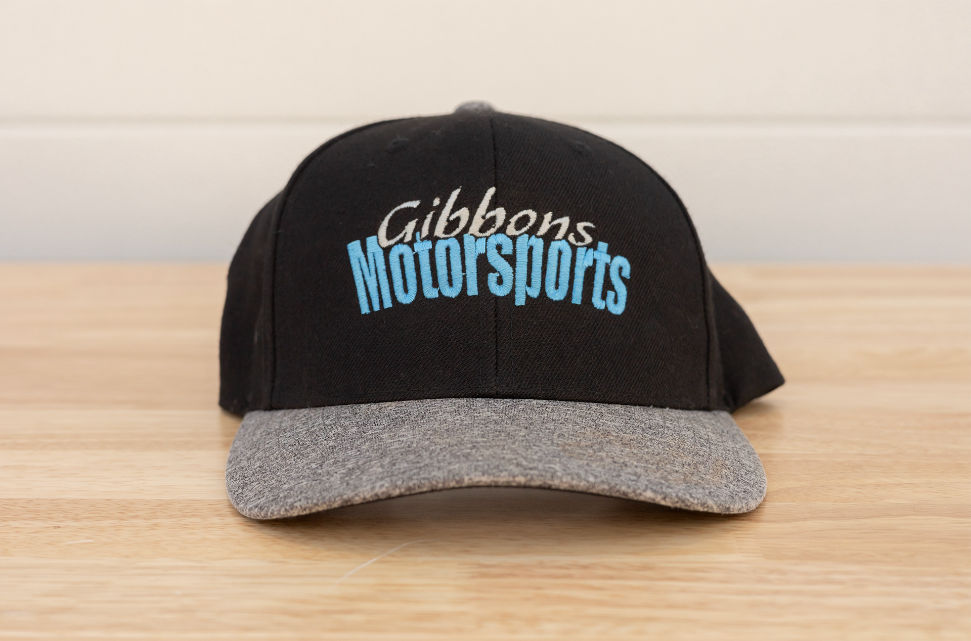 Black Gibbons Motorsports cap with gray brim, sitting on a wooden surface