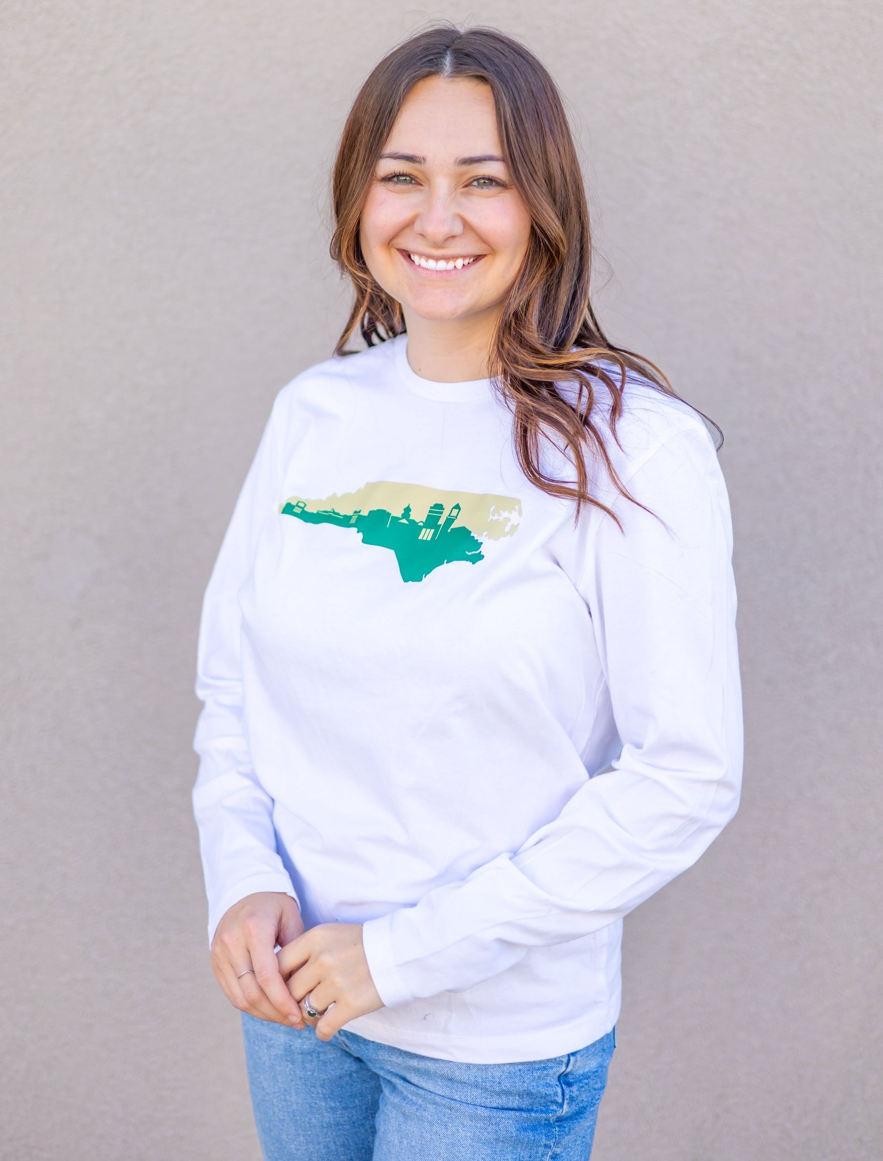 Smiling person in a white long-sleeve shirt with a green graphic, standing against a plain wall.