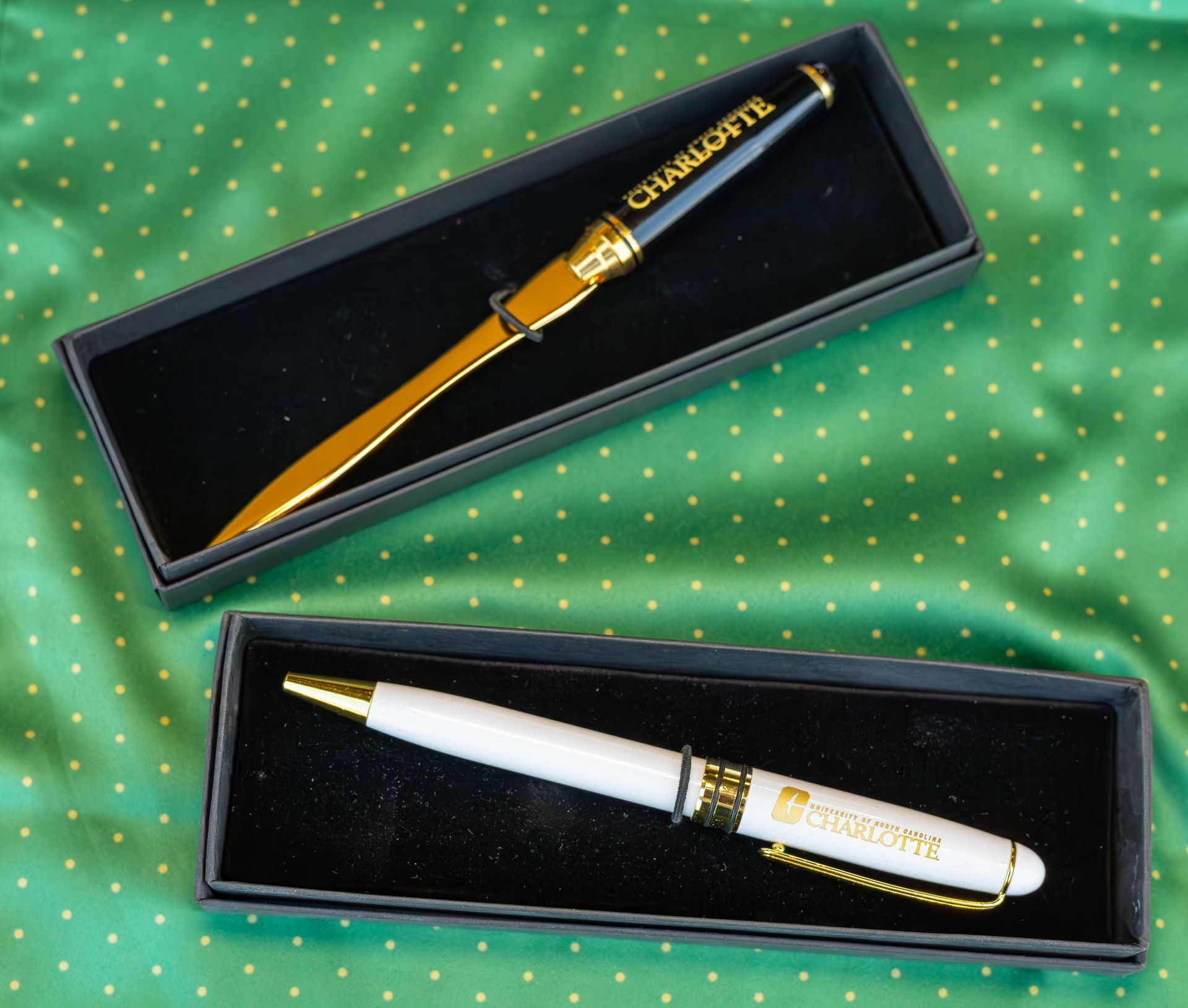 Two gold-and-white pens in black presentation cases on green polka-dot fabric.