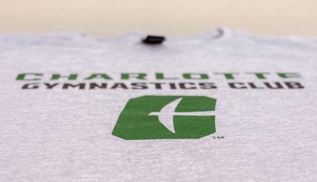 Green-and-black Gymnastics Club logo on a light fabric shirt