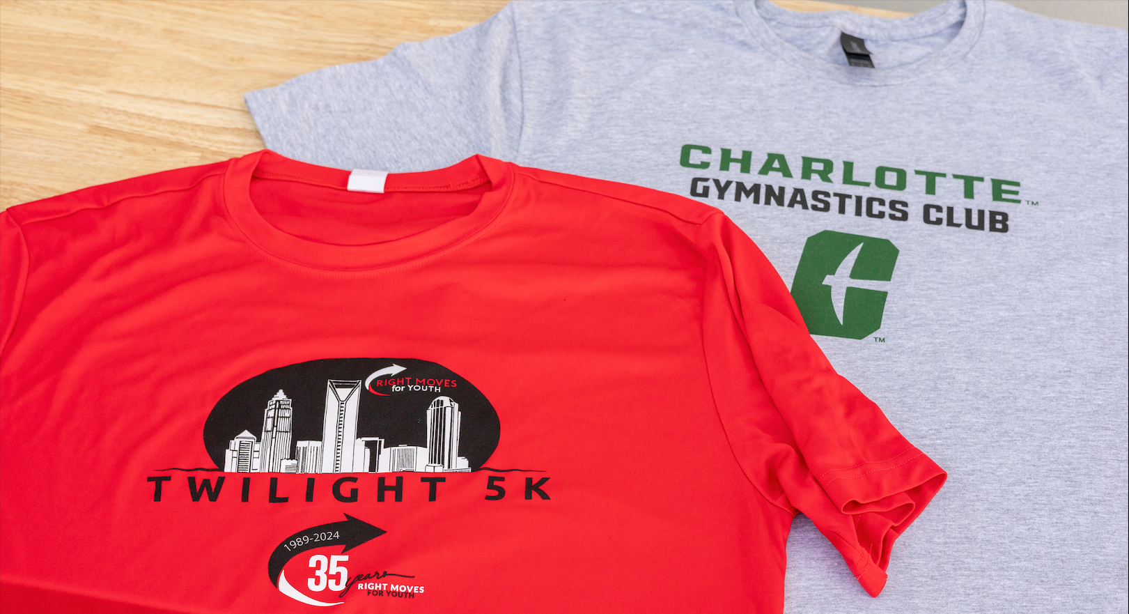 Two T-shirts on a wooden surface: red Twilight 5K shirt and gray Charlotte Gymnastics Club shirt