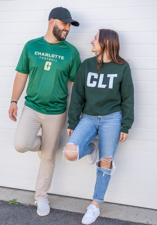Two people in green sweatshirts and jeans leaning against a white wall, smiling at each other.