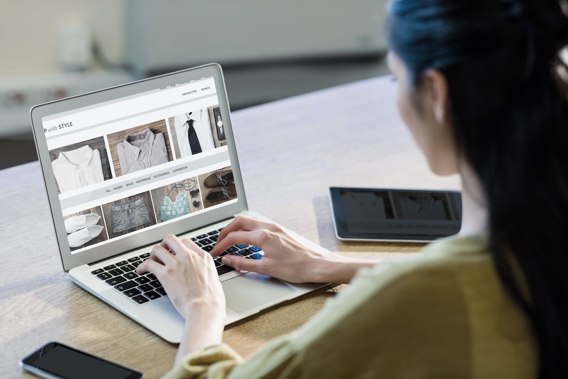 Person browsing clothing on a laptop at a desk, with a phone nearby.