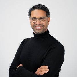 Man met bril, armen over elkaar, glimlachend; draagt ​​een zwarte coltrui.