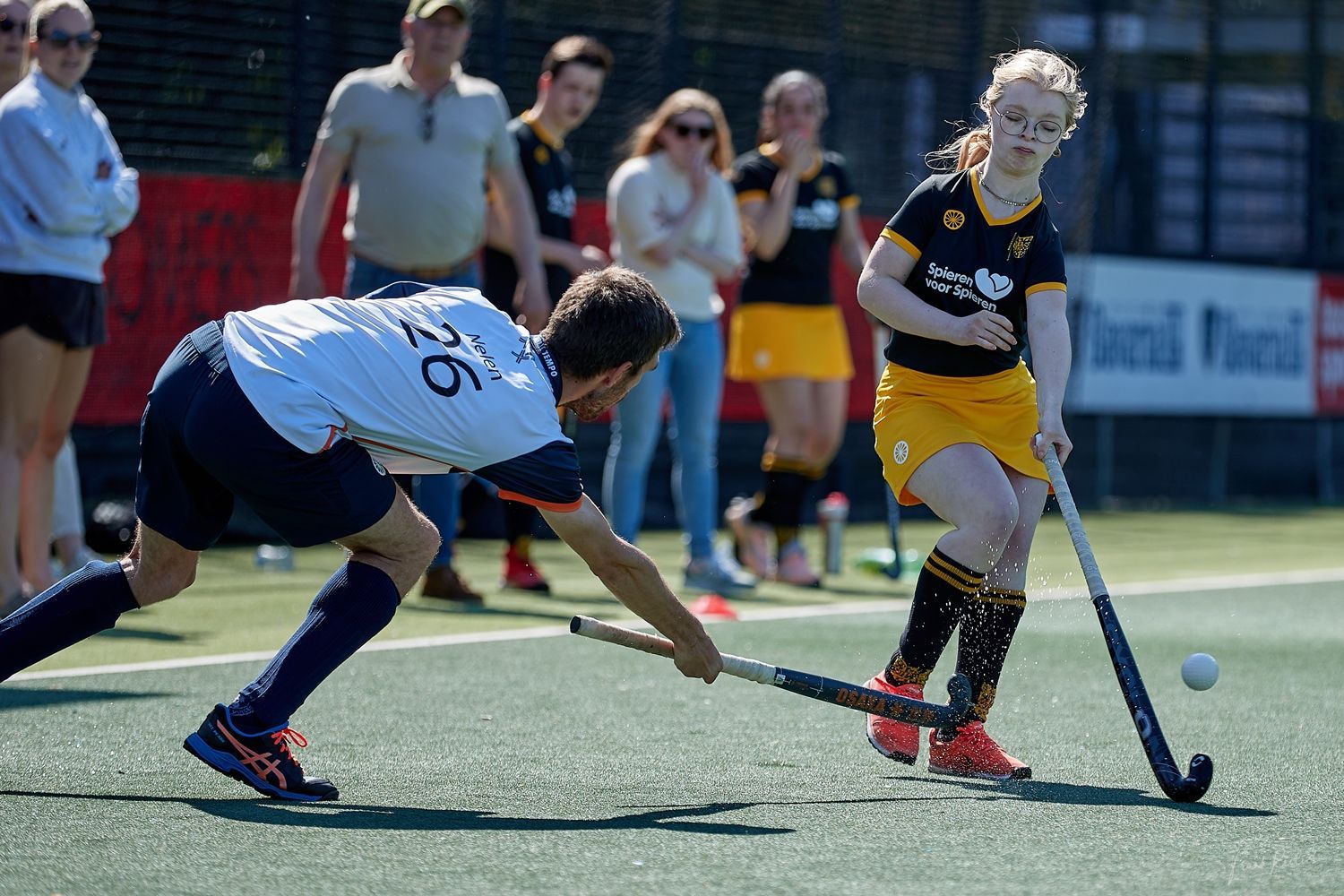 Hockeyspelers op een groen veld. Een speler in het wit duikt naar de bal, terwijl de speler in het zwart zich klaarmaakt om te slaan.