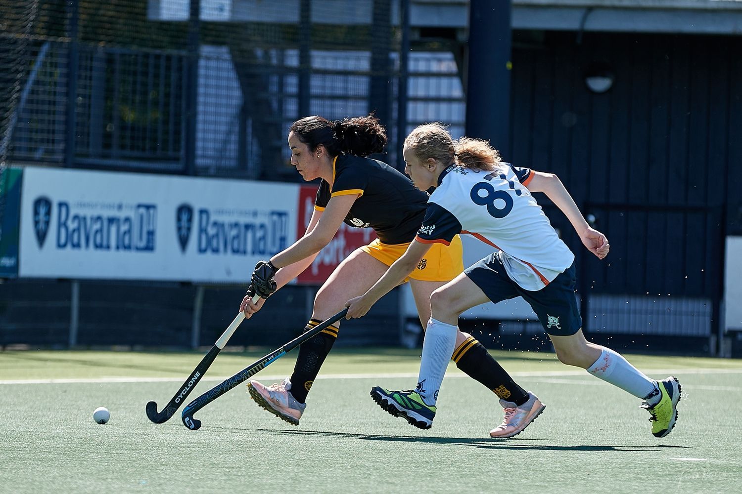 Twee hockeyspelers op een groen veld, de een in zwart, de ander in wit. Ze jagen met sticks achter een bal aan.