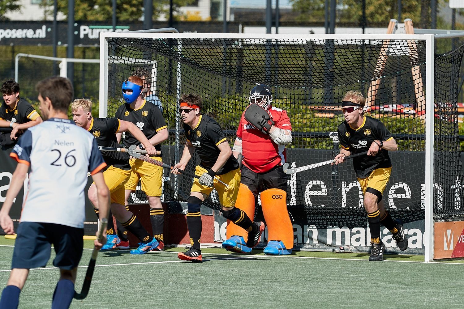 Hockeyspelers bij het doel, in zwart-gele uniformen. Doelman in rood.