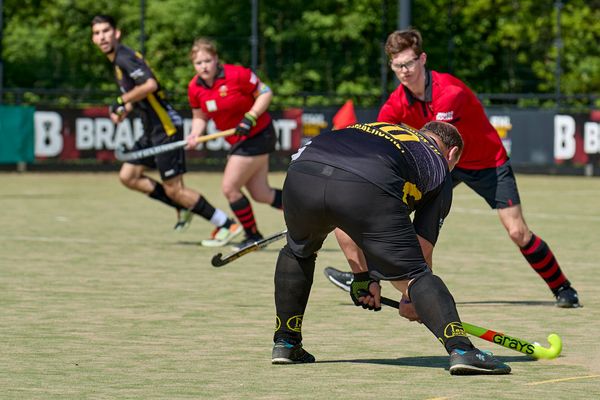 Hockeyspelers in actie, een speler in het zwart tackelt een speler in het rood. Groen veld, fel zonlicht.