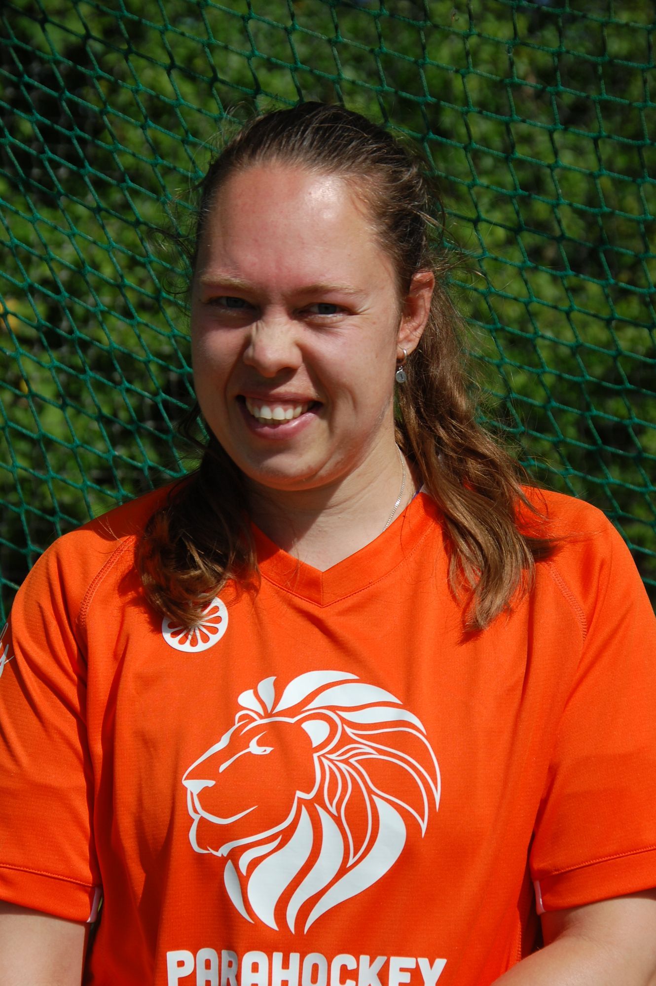 Een lachende vrouw, gekleed in een oranje shirt met een leeuwenlogo, staat voor een groene, doorschijnende achtergrond.