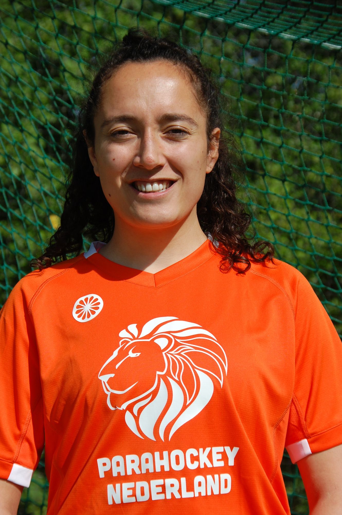 Vrouw in een oranje shirt met een leeuwenlogo, lachend voor een groen net.