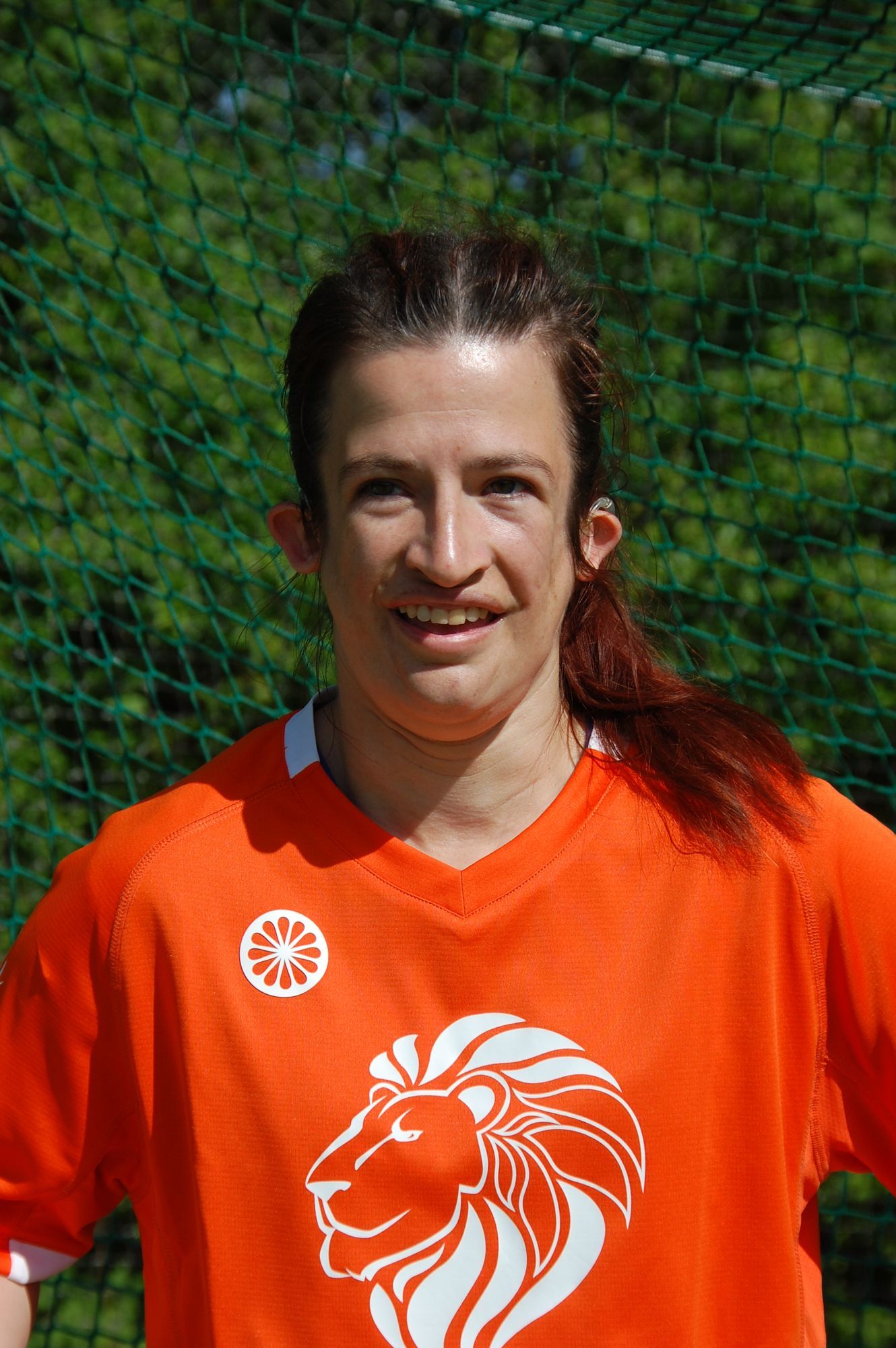 Vrouw in oranje shirt met leeuwenembleem lacht, staand voor een groene achtergrond.