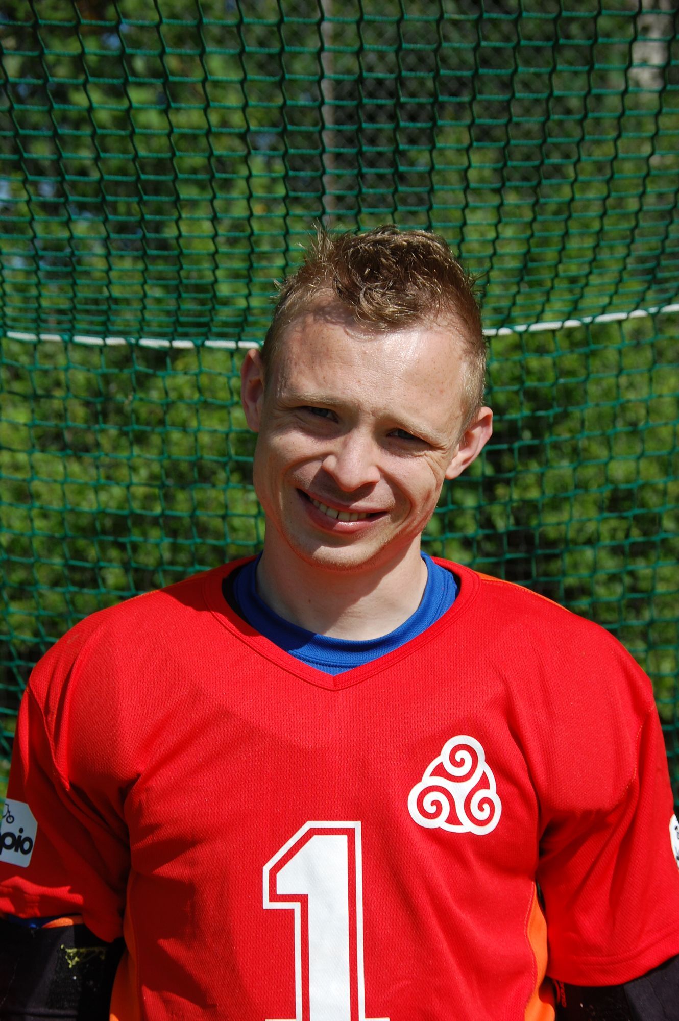 Man in rood shirt met nummer 1, lachend, voor een groene achtergrond van gaas.