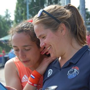 Twee vrouwen glimlachen en kijken naar een telefoon; een van hen heeft een arm om de schouder van de ander geslagen, buiten.