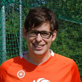 Een man met een bril, gekleed in een oranje shirt, staat lachend naast een groen net.