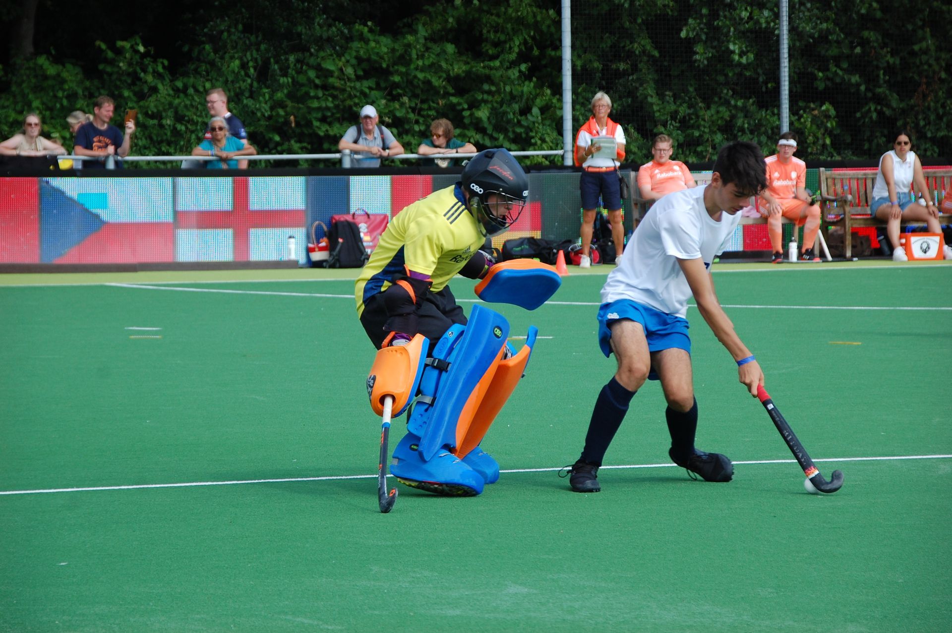 Een hockeyspeler staat tegenover een keeper, beiden op een groen veld. Toeschouwers kijken toe.