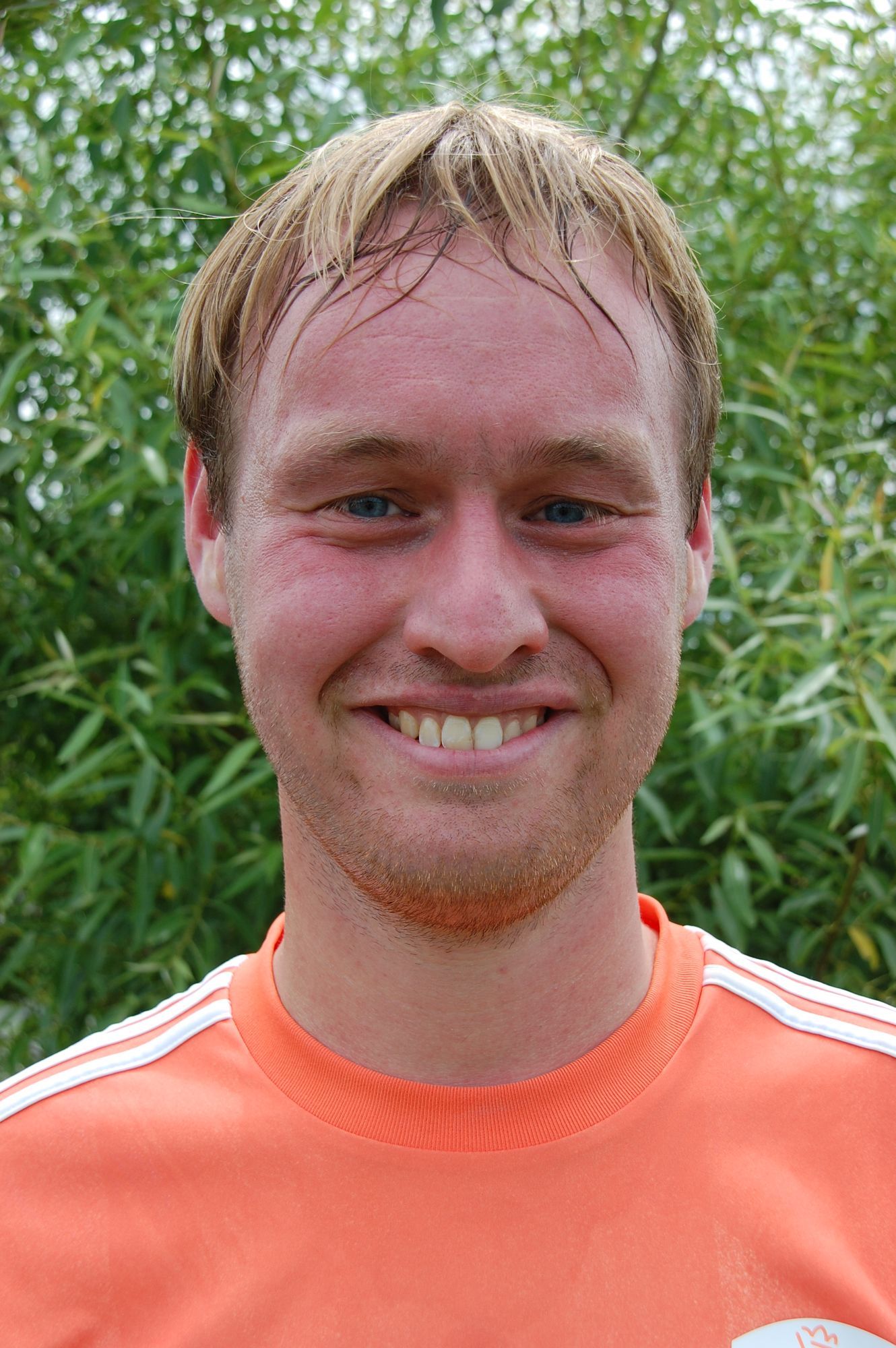 Een man in een oranje shirt lacht naar de camera, met een groene achtergrond.