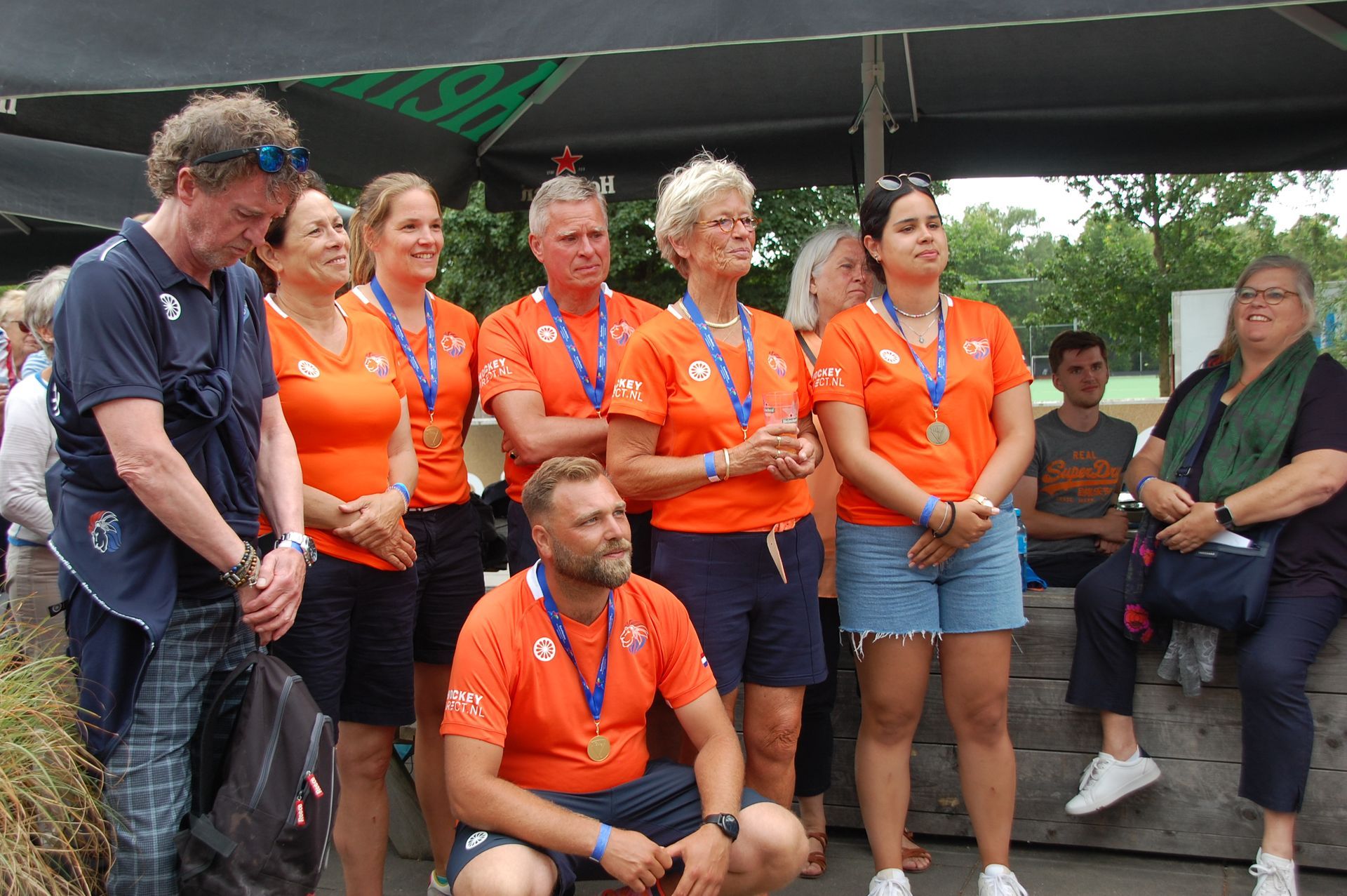 Een groep mensen in oranje shirts, met medailles op, staat buiten. Sommigen glimlachen.