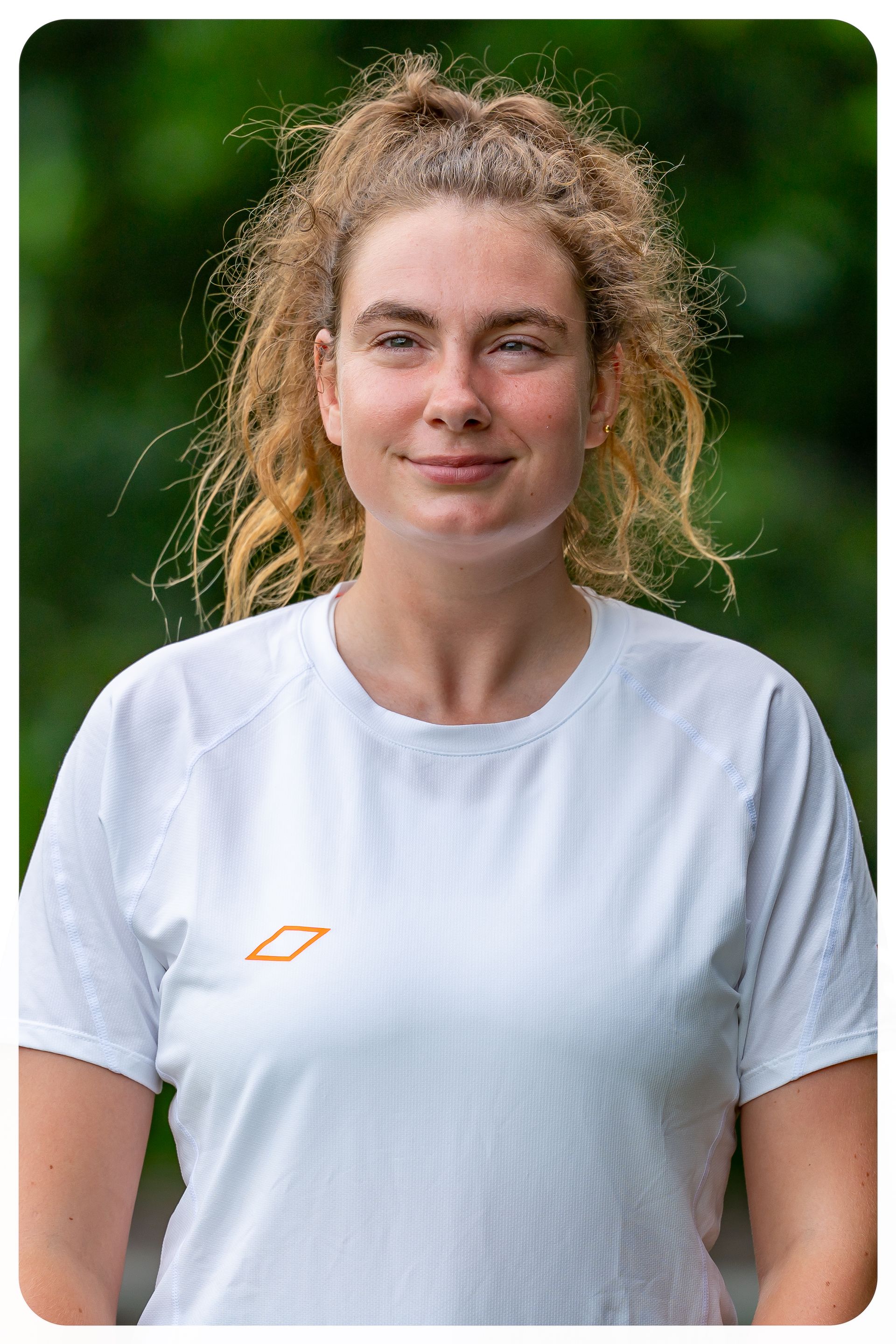Vrouw met krullend blond haar in een witte blouse, die lichtjes glimlacht tegen een wazige groene achtergrond.