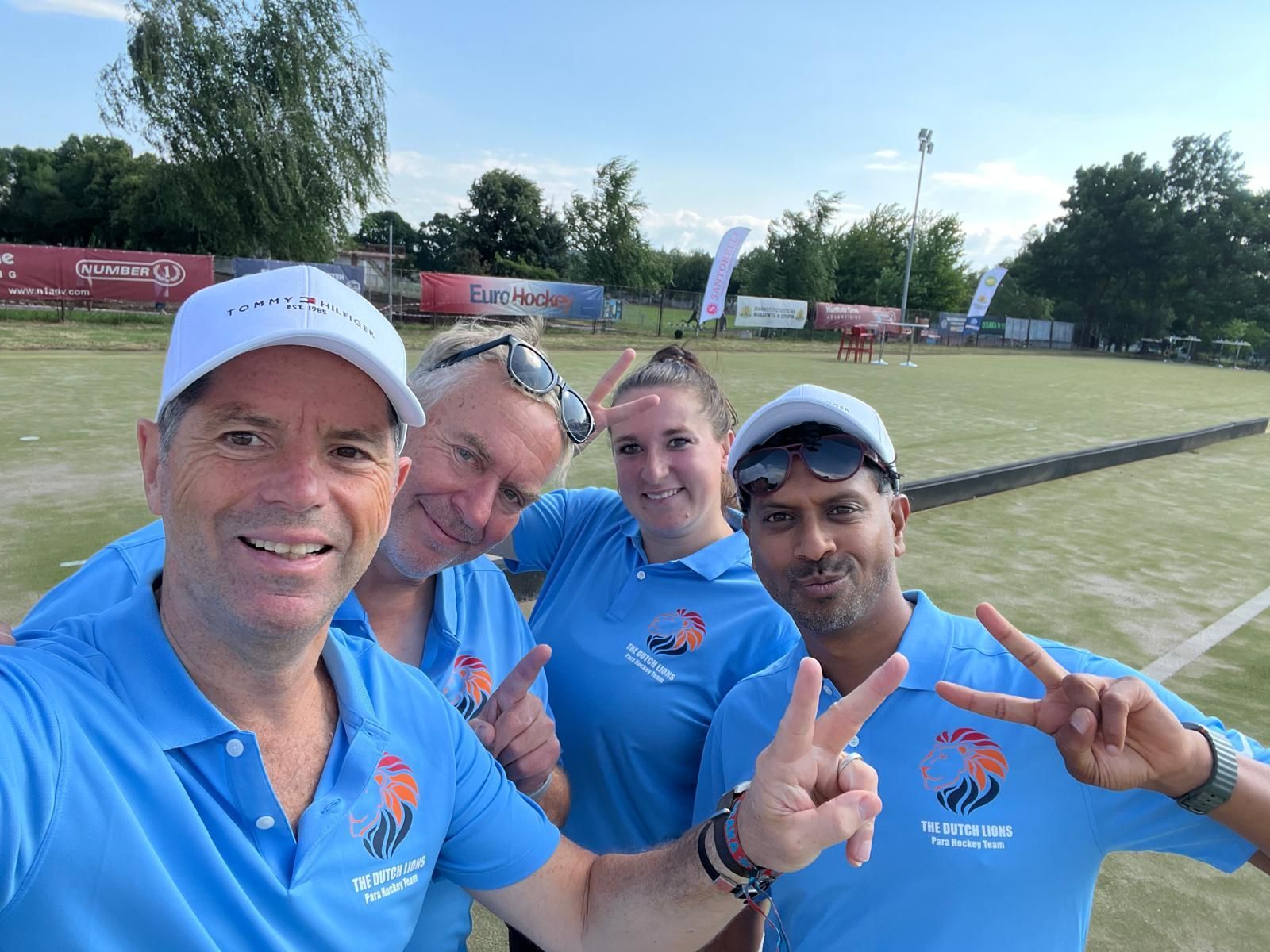 Vier mensen in blauwe shirts en petten poseren voor een selfie op een groen veld en maken daarbij het vredesteken.