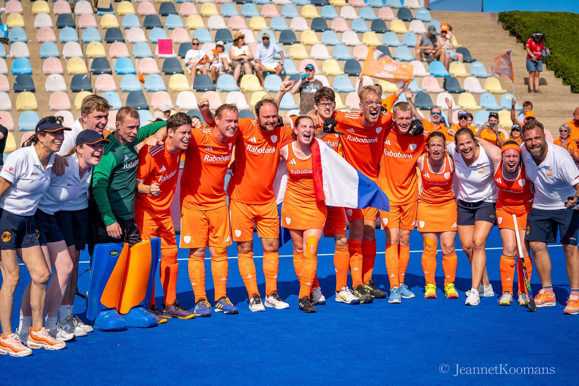 Nederlands hockeyteam in oranje tenues viert de overwinning op een blauw veld en houdt een vlag vast.