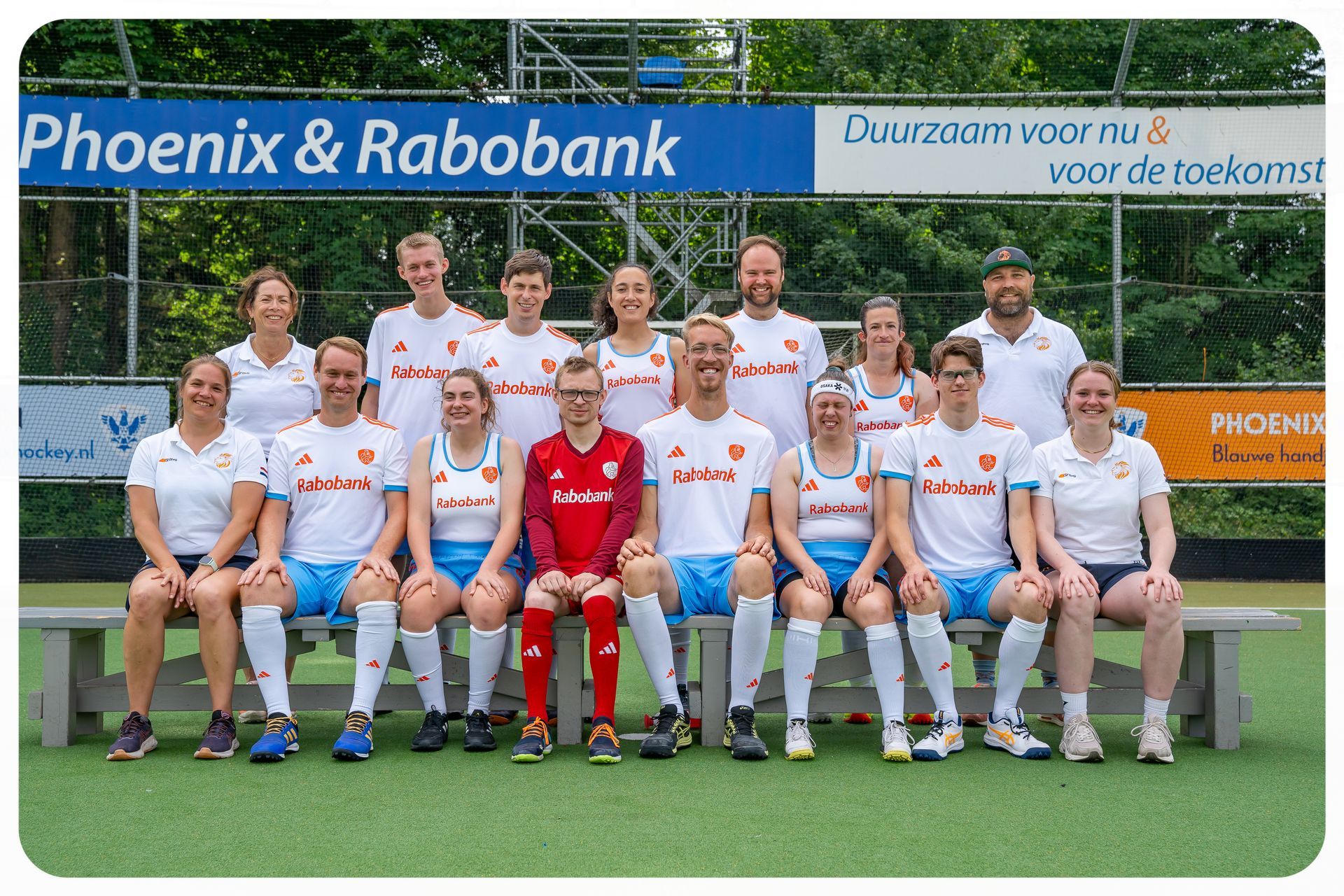 Een hockeyteam poseert voor een spandoek. De spelers dragen witte en oranje uniformen en zitten op een bankje.
