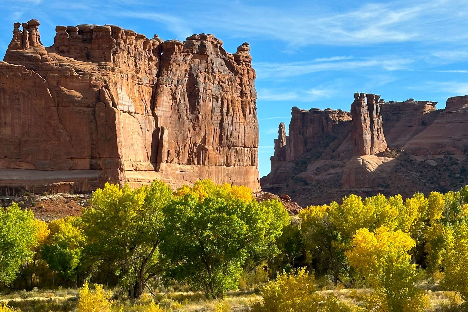 Red rock formations tower above golden trees under a blue sky in a desert landscape.