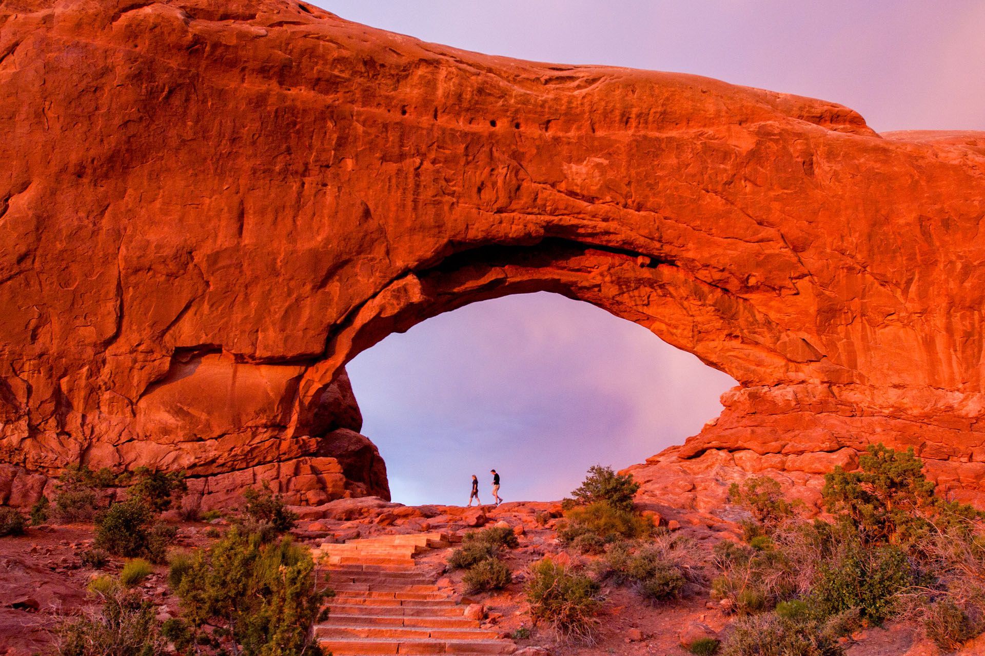 Red rock arch at sunset with two figures standing inside.