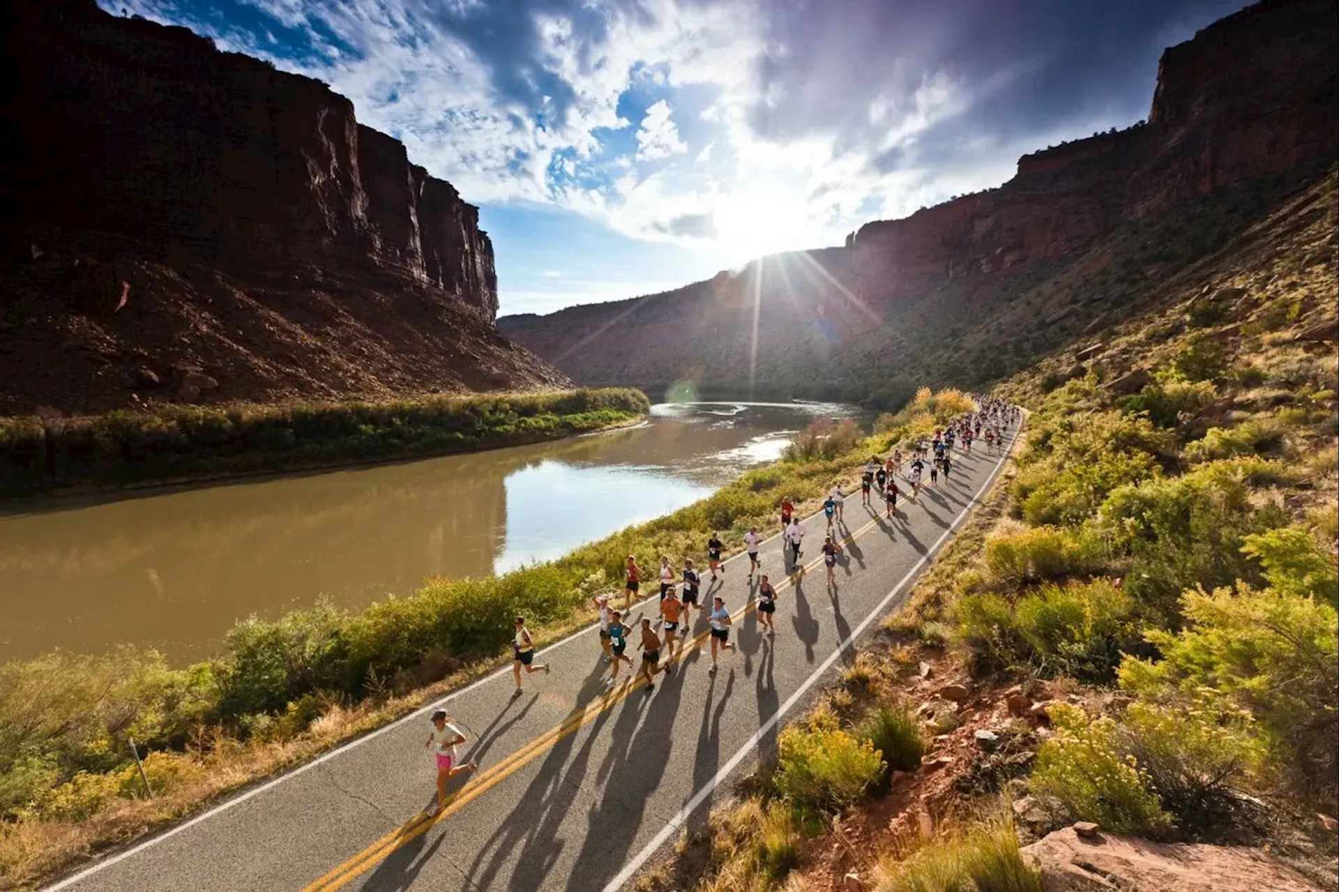 Runners on a road next to a river and canyon, sun shining through the sky.