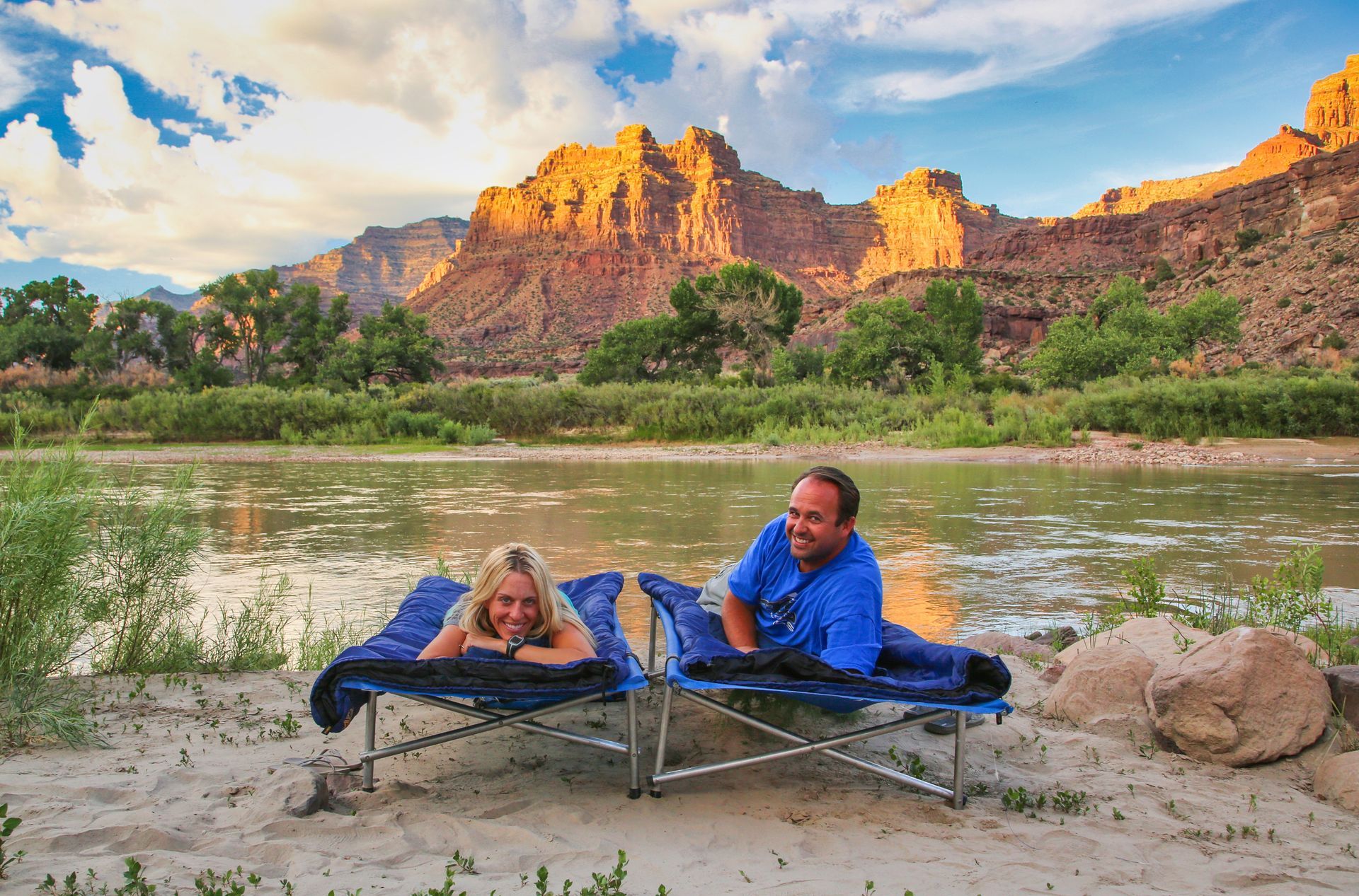 Couple relaxes in camp cots on a sandy riverbank, with a mountain backdrop at sunset.